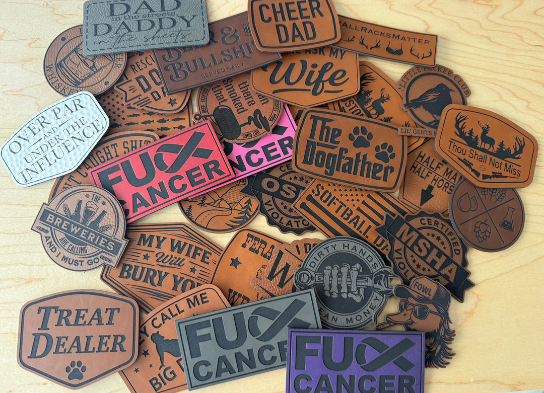 A pile of novelty leather patches with humorous and suggestive slogans, displayed on a light wood background. The patches feature various designs, including brown, pink, purple, and gray tones, with some featuring paw prints and rustic western-style borders.