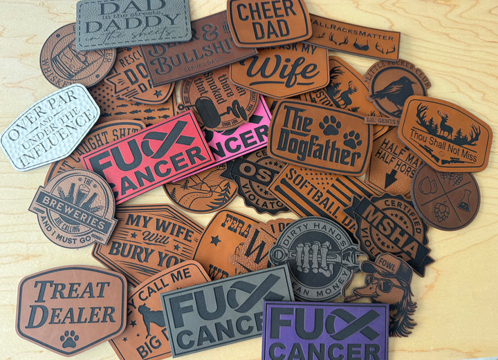A pile of novelty leather patches with humorous and suggestive slogans, displayed on a light wood background. The patches feature various designs, including brown, pink, purple, and gray tones, with some featuring paw prints and rustic western-style borders.