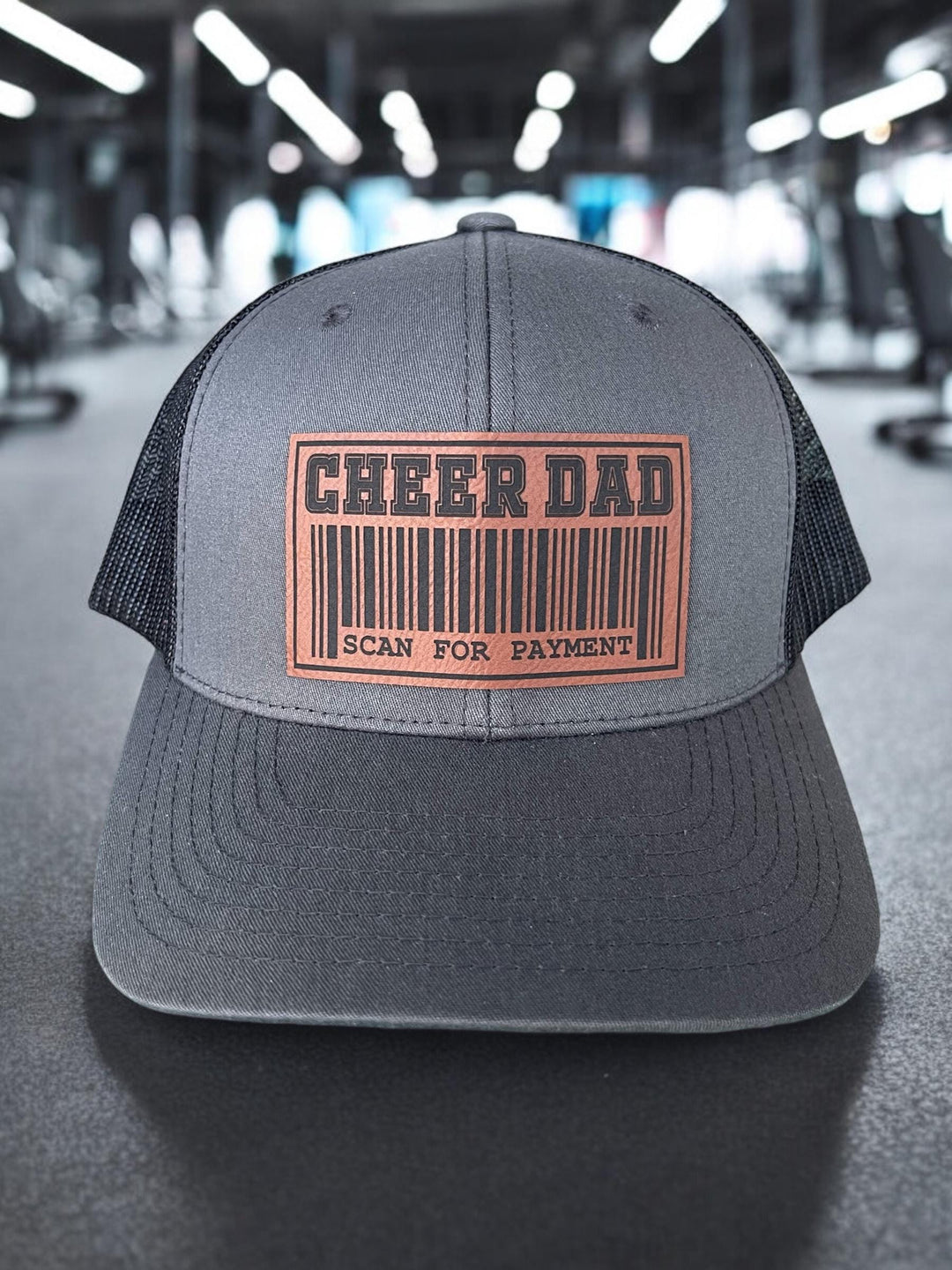 Cheer Dad Trucker Hat with two custom patches: a fun barcode design or a bold &#39;Cheer Dad&#39; text. This hat features a structured charcoal front, breathable black mesh back, and an adjustable snapback for comfort.