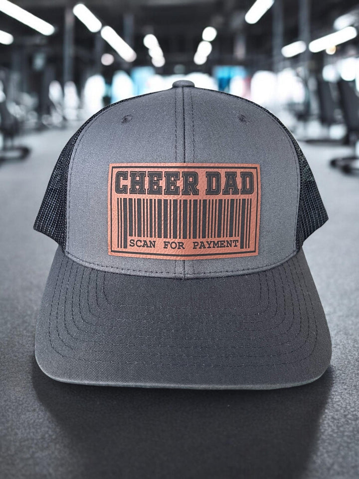 Cheer Dad Trucker Hat with two custom patches: a fun barcode design or a bold &#39;Cheer Dad&#39; text. This hat features a structured charcoal front, breathable black mesh back, and an adjustable snapback for comfort.